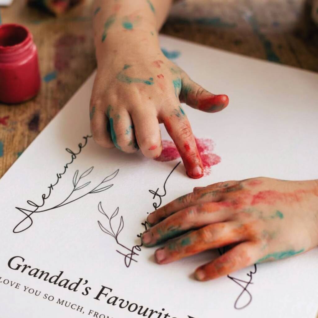 A close-up of two children's hands covered in colorful paint, pressing red and green paint into a personalized white paper craft sheet printed with the name 'Alexander' and 'Grandad's Favourite.