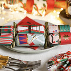 A festive holiday table setting featuring a plate of decorated Christmas crackers with nutcracker and military designs, alongside silver forks and napkins on a white tablecloth with warm blurred lights in the background.