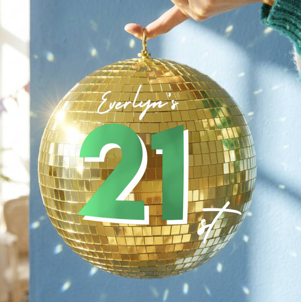 A large gold disco ball with mirrored tiles and a green '21' printed on the front, held by a hand against a blue wall with confetti.