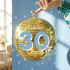 A hand wearing a green and white striped sweater holds a large gold disco ball ornament with the text 'Arabella's 30th' written on it, set against a blue wall with soft light and shadow effects.