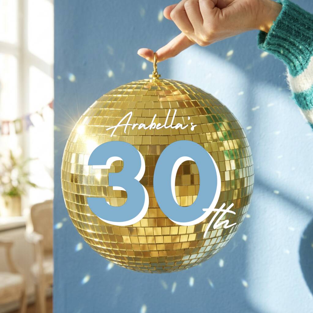 A hand wearing a green and white striped sweater holds a large gold disco ball ornament with the text 'Arabella's 30th' written on it, set against a blue wall with soft light and shadow effects.