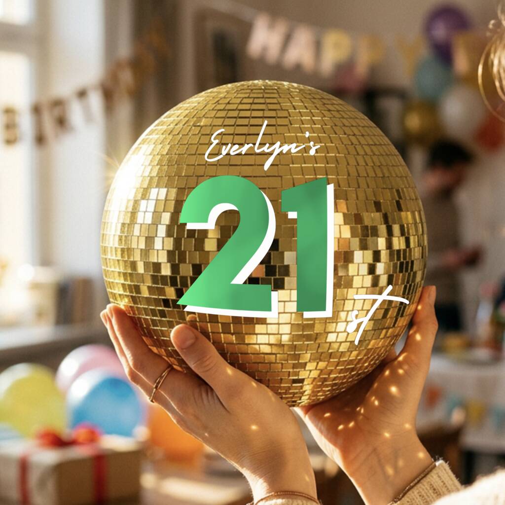 A person holds a gold disco ball party decoration in both hands, featuring a green and white graphic that reads 'Evelyn's 21st'. The background shows a softly blurred birthday party setting with balloons and a 'HAPPY BIRTHDAY' banner.