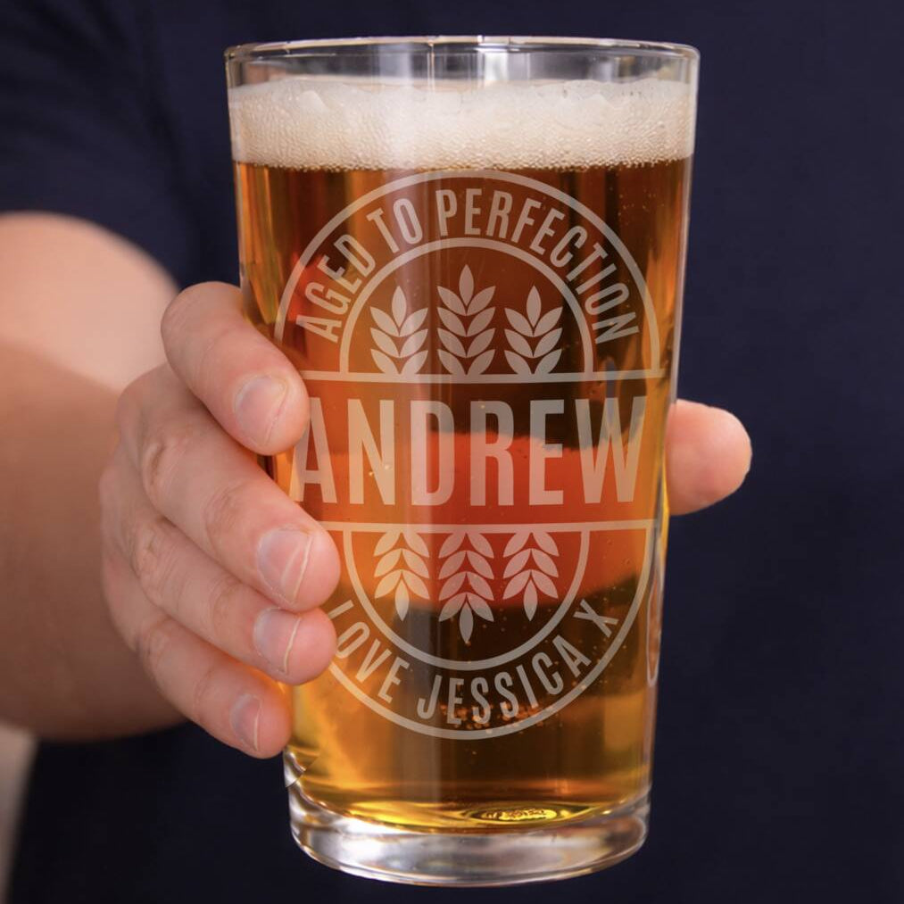 A hand holding a clear pint glass filled with amber beer, featuring a frosted custom engraving that reads 'AGED TO PERFECTION', 'ANDREW', and 'LOVE JESSICA X' with wheat motifs. The background shows a person's hand and dark navy shirt.