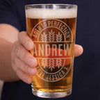 A hand holding a clear pint glass filled with amber beer, featuring a frosted custom engraving that reads 'AGED TO PERFECTION', 'ANDREW', and 'LOVE JESSICA X' with wheat motifs. The background shows a person's hand and dark navy shirt.