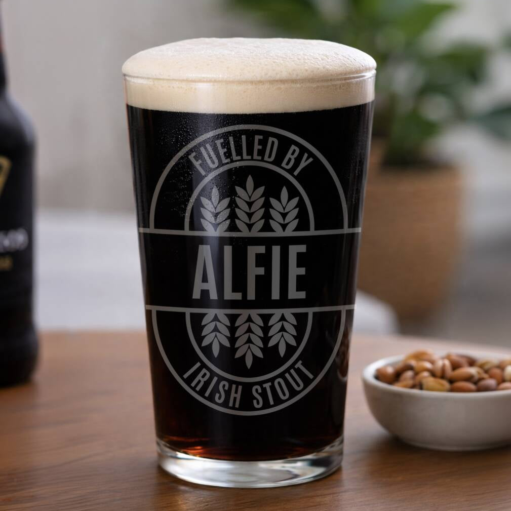 A pint glass filled with dark stout beer, topped with a thick white foam head, standing on a wooden table. The glass has a circular gray graphic that reads 'FUELLED BY ALFIE IRISH STOUT' with wheat illustrations.