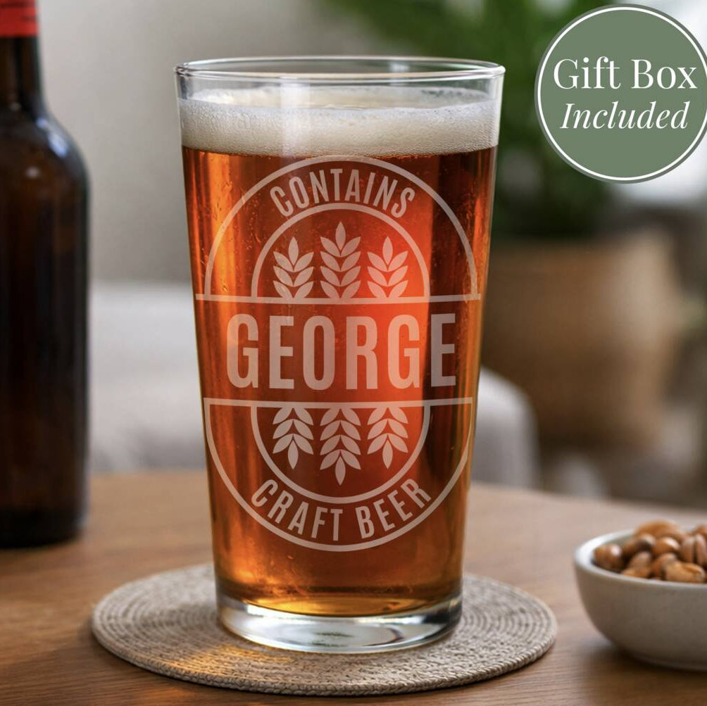 A personalized clear glass beer tumbler filled with amber beer, shown on a woven coaster in a real indoor setting with a bottle, bowl of nuts, and a green 'Gift Box Included' badge. The glass has a frosted white circular design with wheat motifs and the name 'GEORGE'.