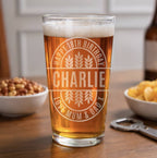 A clear pint glass filled with amber beer stands on a wooden table, etched with a circular design reading 'HAPPY 18TH BIRTHDAY CHARLIE' and 'JUNE MUM & DAD' with wheat motifs. Bowls of snacks and a bottle are blurred in the background.