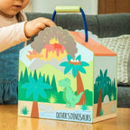 A child's hand is holding a colorful cardboard dinosaur-themed gift box with a blue handle. The box features a prehistoric scene with a green T-Rex, palm trees, mountains, and an erupting volcano, with the text 'OLIVER'S DINOSAURS' printed on the front.