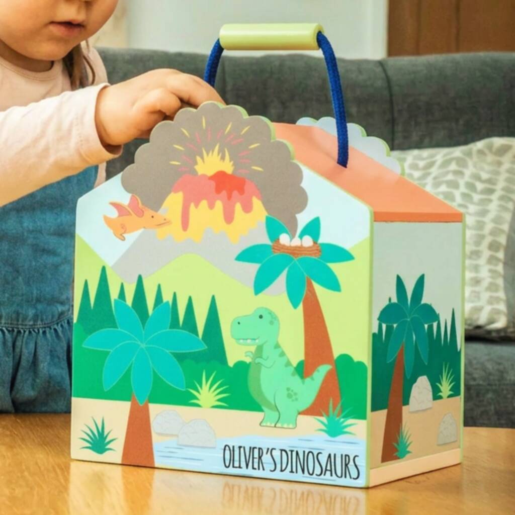 A child's hand is holding a colorful cardboard dinosaur-themed gift box with a blue handle. The box features a prehistoric scene with a green T-Rex, palm trees, mountains, and an erupting volcano, with the text 'OLIVER'S DINOSAURS' printed on the front.