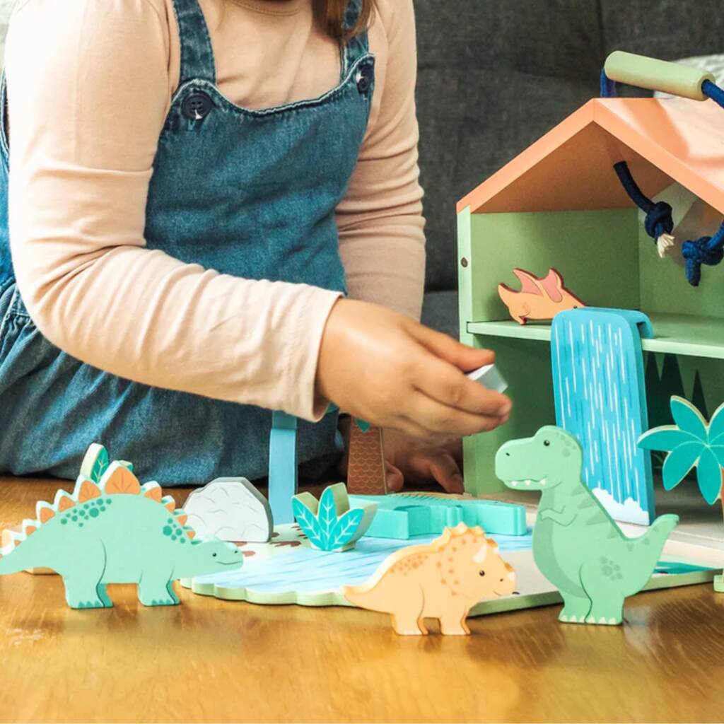 A child wearing denim overalls and a beige long-sleeve shirt plays with a wooden dinosaur and house toy set on a wooden table.