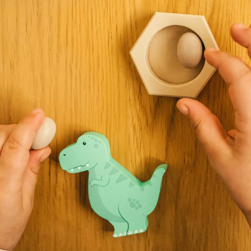 A child's hands interacting with a wooden dinosaur nesting egg toy on a warm wood-grain tabletop. The mint green dinosaur piece is on the floor, while one hand holds a small white egg and the other hand is placing another egg into a hexagonal wooden holder.