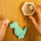 A child's hands interacting with a wooden dinosaur nesting egg toy on a warm wood-grain tabletop. The mint green dinosaur piece is on the floor, while one hand holds a small white egg and the other hand is placing another egg into a hexagonal wooden holder.