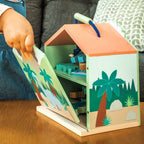 A child's hand is opening a wooden toy house box on a wooden table, with a gray couch in the background. The toy house has a peach-colored roof, a blue handle, and colorful tropical jungle illustrations on the front and inside.