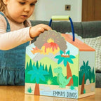 A young child in blue denim overalls sits at a wooden table, holding the handle of a colorful dinosaur-themed gift box. The box is printed with illustrations of a green T-Rex, palm trees, a volcano eruption, and the personalized text 'EMMA'S DINOS'.