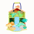A wooden dinosaur playset in the shape of a house with a blue handle, featuring colorful dinosaur figures, a waterfall, palm trees, and a sandy ground on a white background.