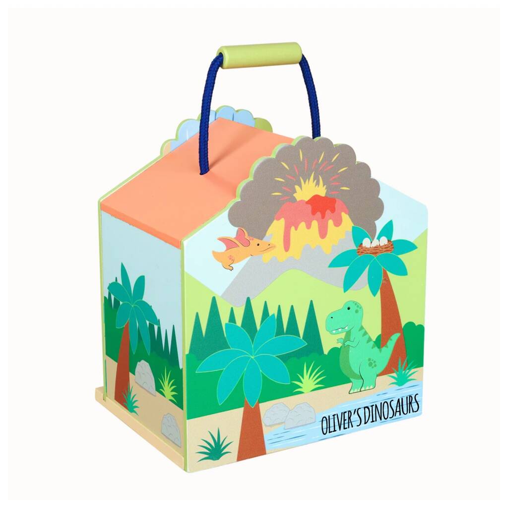 A colorful wooden house-shaped children's toy box with a dinosaur theme, featuring an illustrated prehistoric landscape with palm trees, a green T-Rex, an erupting volcano, and the text 'Oliver's Dinosaur'. It has a light green handle and a blue rope handle, shown on a plain white background.