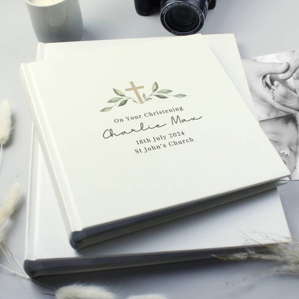 A personalized baby christening photo album with a cream cover, featuring a gold cross and green leaf design, styled on a light gray surface with a camera, candle, and baby photo in the background.