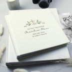 A personalized baby christening photo album with a cream cover, featuring a gold cross and green leaf design, styled on a light gray surface with a camera, candle, and baby photo in the background.