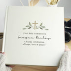 A personalized hardcover photo album held by hands in a cozy indoor setting. The album cover is a warm cream color with a printed olive green cross and leaf design, and custom black text reading 'First Holy Communion', 'Imogen Bailey', and 'A happy celebration of hope, love & prayer'.