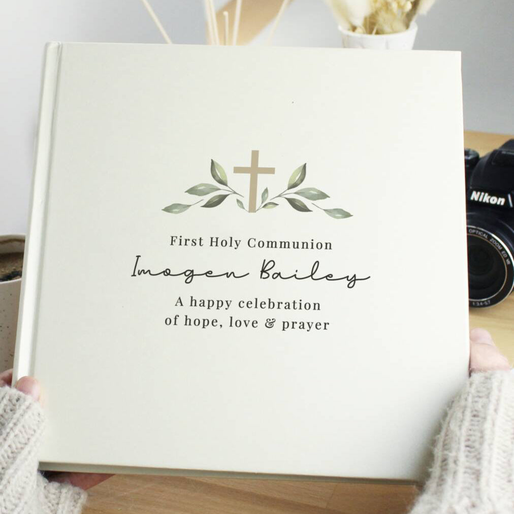 A personalized hardcover photo album held by hands in a cozy indoor setting. The album cover is a warm cream color with a printed olive green cross and leaf design, and custom black text reading 'First Holy Communion', 'Imogen Bailey', and 'A happy celebration of hope, love & prayer'.