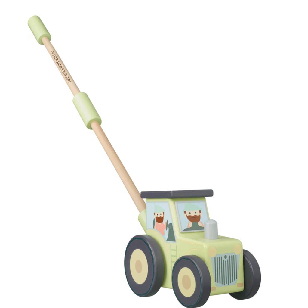 A wooden push toy tractor with a light green body, dark gray wheels, and a natural wood handle with light green grips. The windows show cartoon driver figures.
