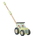 A wooden push toy tractor with a light green body, dark gray wheels, and a natural wood handle with light green grips. The windows show cartoon driver figures.