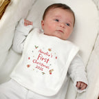 A baby wearing a white long-sleeve bodysuit and bib, lying on a white quilted bassinet. The bib is personalized with the text 'Amelia's First Christmas 2026' in red, decorated with small festive illustrations.