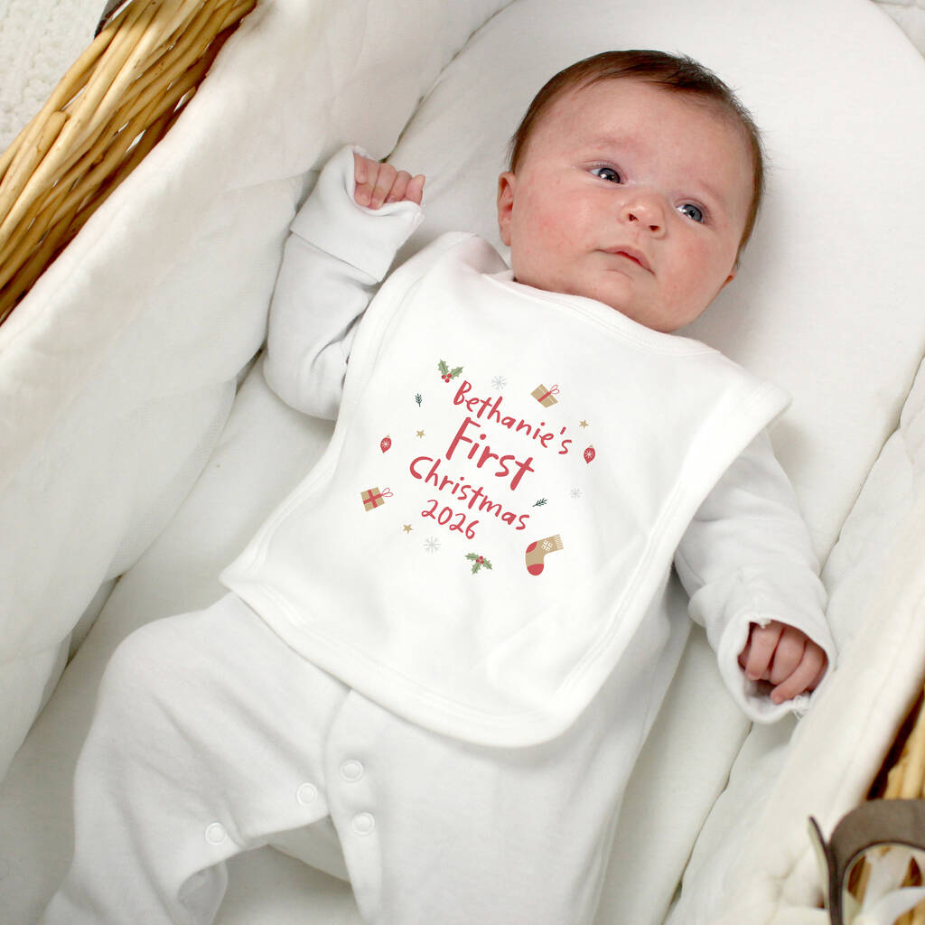 A baby lying in a wicker bassinet wearing a white long-sleeve bodysuit and bib. The bib has a red and pink printed design that reads 'Bethanie's First Christmas 2026' with small holiday illustrations.