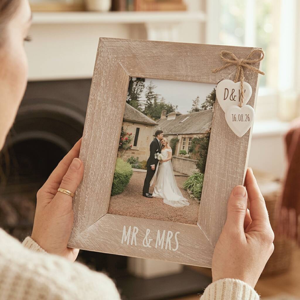 A person holds a rustic light wood photo frame with 'MR & MRS' and 'D&H' printed on it, featuring a small heart-shaped tag tied with twine, in a cozy indoor setting.