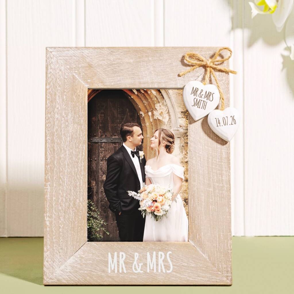 A rustic light wood photo frame with 'MR & MRS' printed on the bottom, holding a wedding photograph of a couple. The frame is decorated with a small twine bow and two heart-shaped tags, and is displayed on a green surface against a white paneled background.