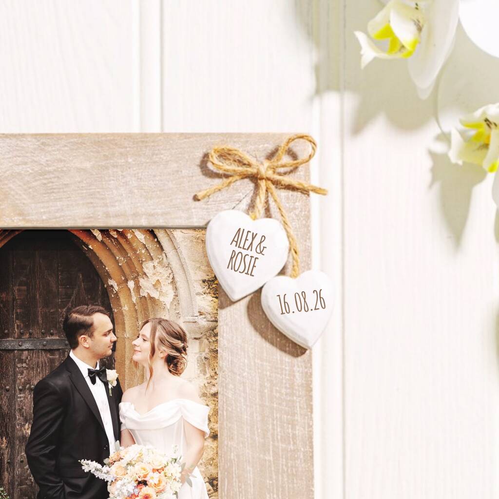 A rustic wooden photo frame with a natural finish, featuring two small white heart-shaped hanging tags tied with twine. One tag is personalized with the names 'ALEX & ROSIE' and the other with the date '16.