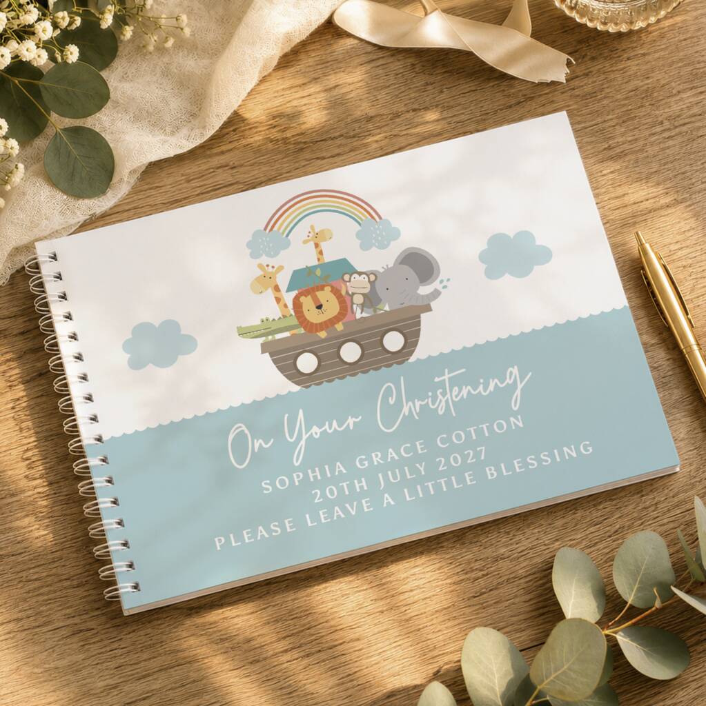 A personalized spiral-bound baby shower game board or notebook lies on a wooden tabletop, decorated with eucalyptus leaves, a gold pen, and a cream ribbon. The cover features a Noah's Ark illustration with a rainbow and clouds, and custom text reading 'On Your Christening' with the name 'Sophia Grace Cotton' and a date.
