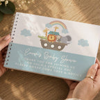 A spiral-bound baby shower gift basket or box cover held by two hands over a wooden tabletop. The cover features a white top half with a colorful Noah's Ark animal illustration and a light blue bottom half with the text 'Emma's Baby Shower' and a thank-you message.