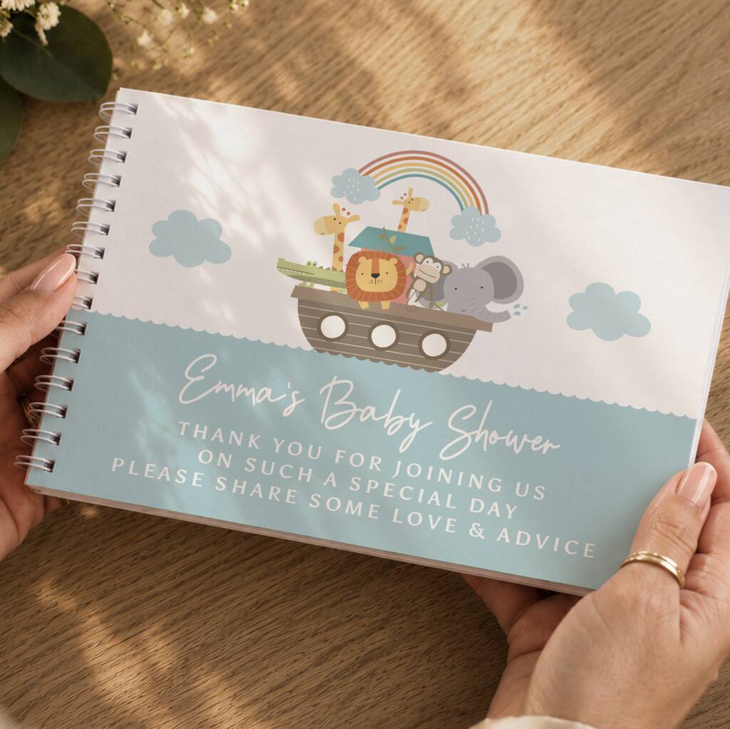 A spiral-bound baby shower gift basket or box cover held by two hands over a wooden tabletop. The cover features a white top half with a colorful Noah's Ark animal illustration and a light blue bottom half with the text 'Emma's Baby Shower' and a thank-you message.