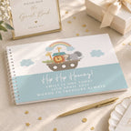 A spiral-bound baby shower guest book with a Noah's Ark illustration and the text 'Hip Hip Hooray! Emily is one today 12th April 2027 Words to treasure always' is displayed on a beige fabric surface with gold confetti, a gold pen, a framed guest book sign, and a gift box.