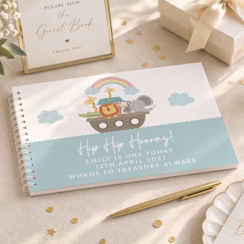 A spiral-bound baby shower guest book with a Noah's Ark illustration and the text 'Hip Hip Hooray! Emily is one today 12th April 2027 Words to treasure always' is displayed on a beige fabric surface with gold confetti, a gold pen, a framed guest book sign, and a gift box.