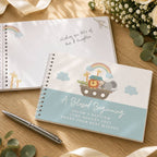A personalized baby baptism keepsake notebook with a spiral binding, featuring a Noah's Ark illustration and the text 'A Blessed Beginning' on a light blue cover. Another open notebook showing a rainbow and giraffe is in the background, styled on a wooden surface with a silver pen and baby flowers.