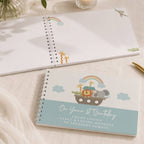 Two spiral-bound baby milestone notebooks on a beige fabric surface. The top notebook is open, showing blank white pages with small safari animal illustrations.
