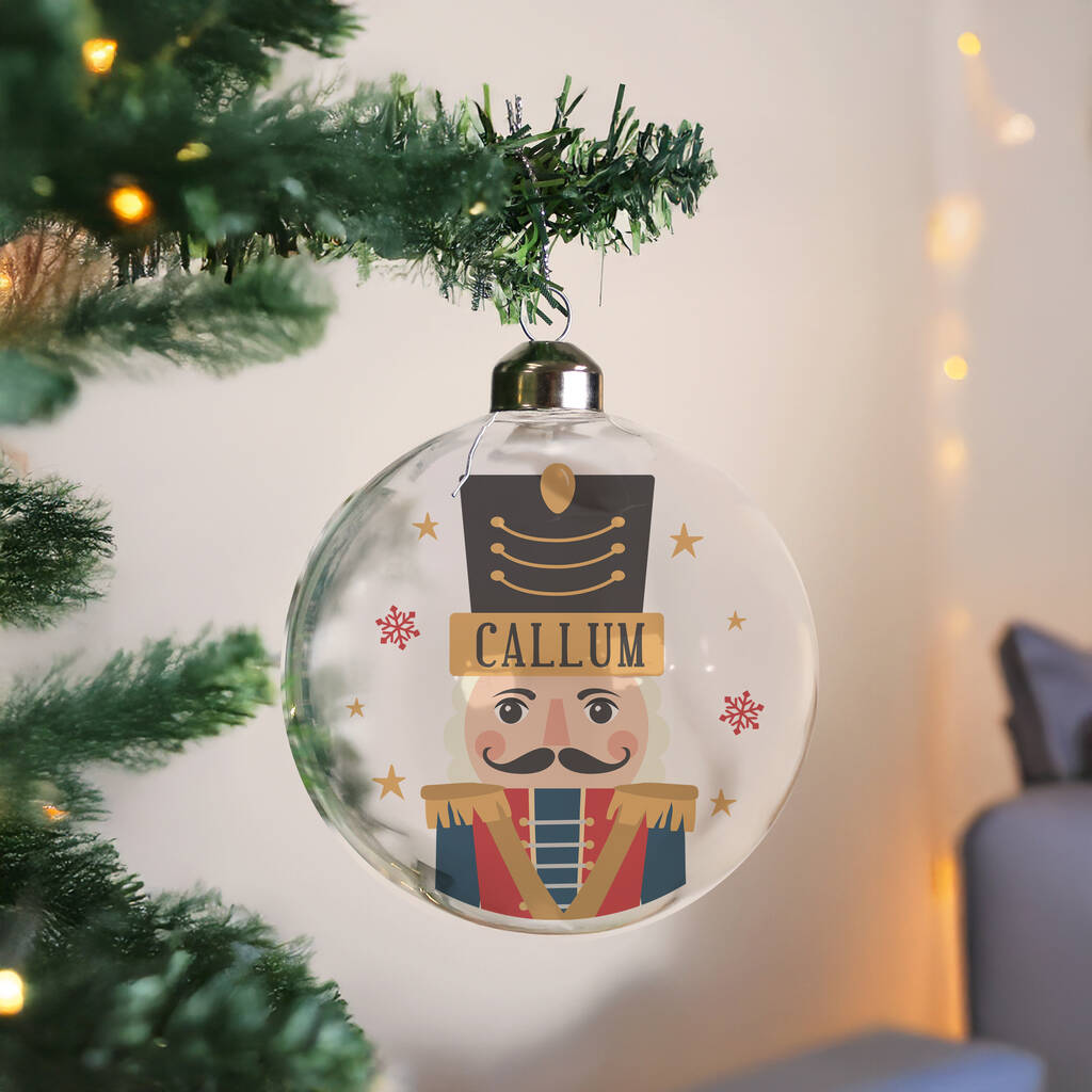 A personalized clear glass Christmas ornament shaped like a nutcracker soldier, hanging from a green tinsel Christmas tree branch. The ornament features a printed face, a black hat with gold stripes, and the name 'CALLUM' on a gold banner.