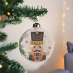 A personalized clear glass Christmas ornament shaped like a nutcracker soldier, hanging from a green tinsel Christmas tree branch. The ornament features a printed face, a black hat with gold stripes, and the name 'CALLUM' on a gold banner.
