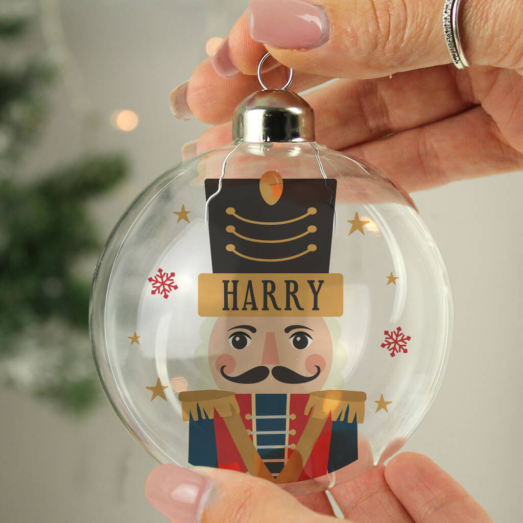 A clear glass Christmas ornament held by hands, featuring a printed nutcracker design with the name 'HARRY' on a gold banner and small red snowflakes.