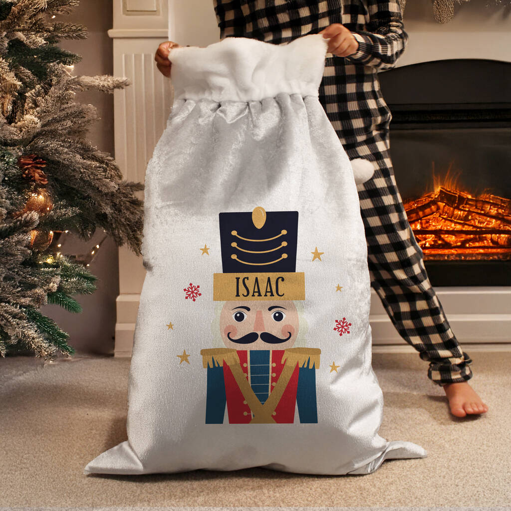 A person in black-and-white checkered pajamas holds a large white Christmas stocking-style sack with a printed nutcracker design. The sack is personalized with the name ISAAC on a gold banner.