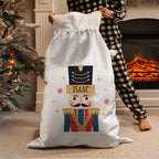 A person in black-and-white checkered pajamas holds a large white Christmas stocking-style sack with a printed nutcracker design. The sack is personalized with the name ISAAC on a gold banner.