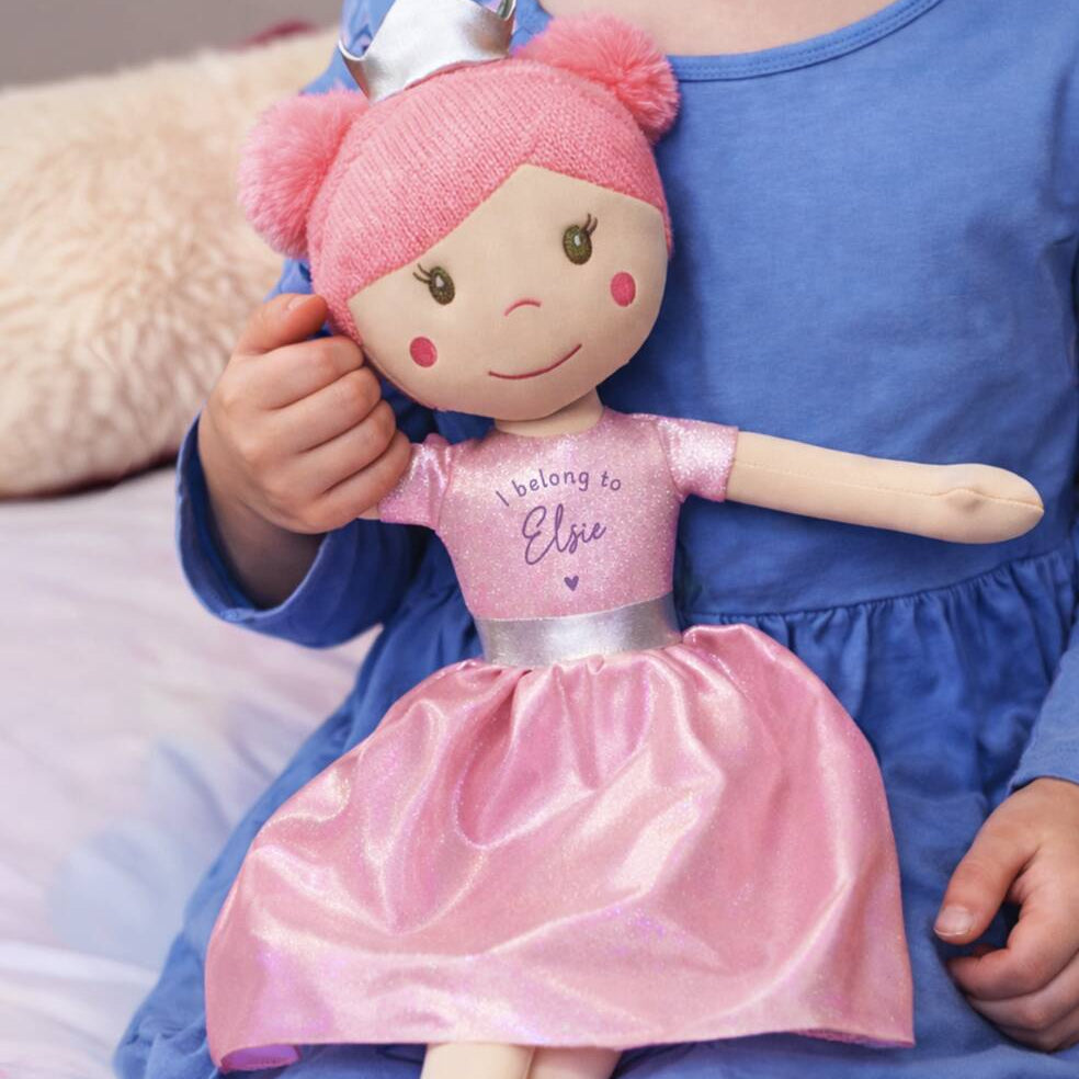 A close-up of a child in a blue dress holding a pink plush doll with a white crown and a shiny pink dress that reads 'I belong to Elise'.