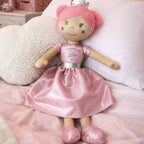 A pink and beige wooden doll with a shiny pink dress, pink hair, and a silver crown, lying on a pink bed with white pillows.