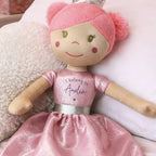 A custom plush doll with a pink knit hat, beige fabric skin, and a glittery pink dress that reads 'I belong to Amelia' on the chest, resting on a white textured pillow.