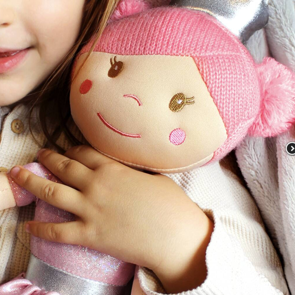 A close-up lifestyle shot of a young child hugging a pink knitted doll with embroidered facial features and a silver bow, wearing a cream-colored knit sweater.