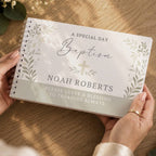 A personalized spiral-bound baptism keepsake book held by two hands over a wooden table. The book cover is white with elegant gray botanical leaf designs, a central gold cross, and personalized text reading 'A SPECIAL DAY Baptism NOAH ROBERTS'.