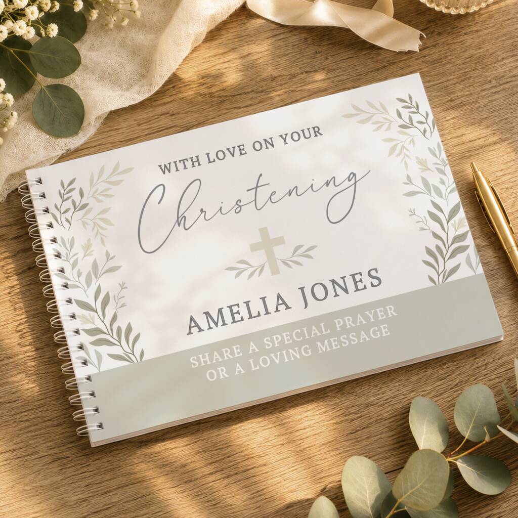 A personalized spiral-bound christening journal lies open on a wooden tabletop, surrounded by eucalyptus leaves, a cream ribbon, a gold pen, and a beige fabric. The cover reads 'WITH LOVE ON YOUR Christening AMELIA JONES' with a small cross and leafy border.