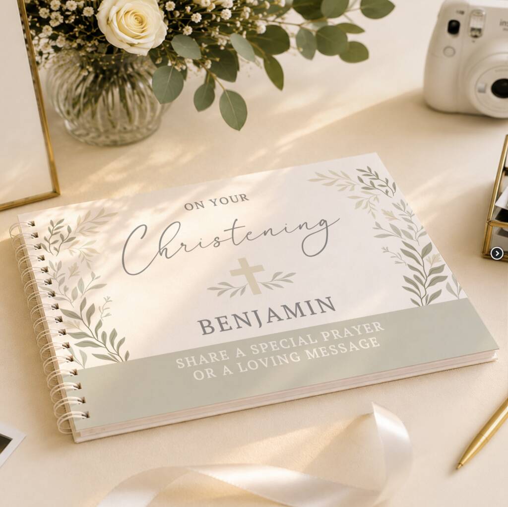 A personalized spiral-bound Christening book or journal lies open on a light beige surface. The cover reads 'ON YOUR Christening BENJAMIN' with floral illustrations and a pale green band at the bottom.